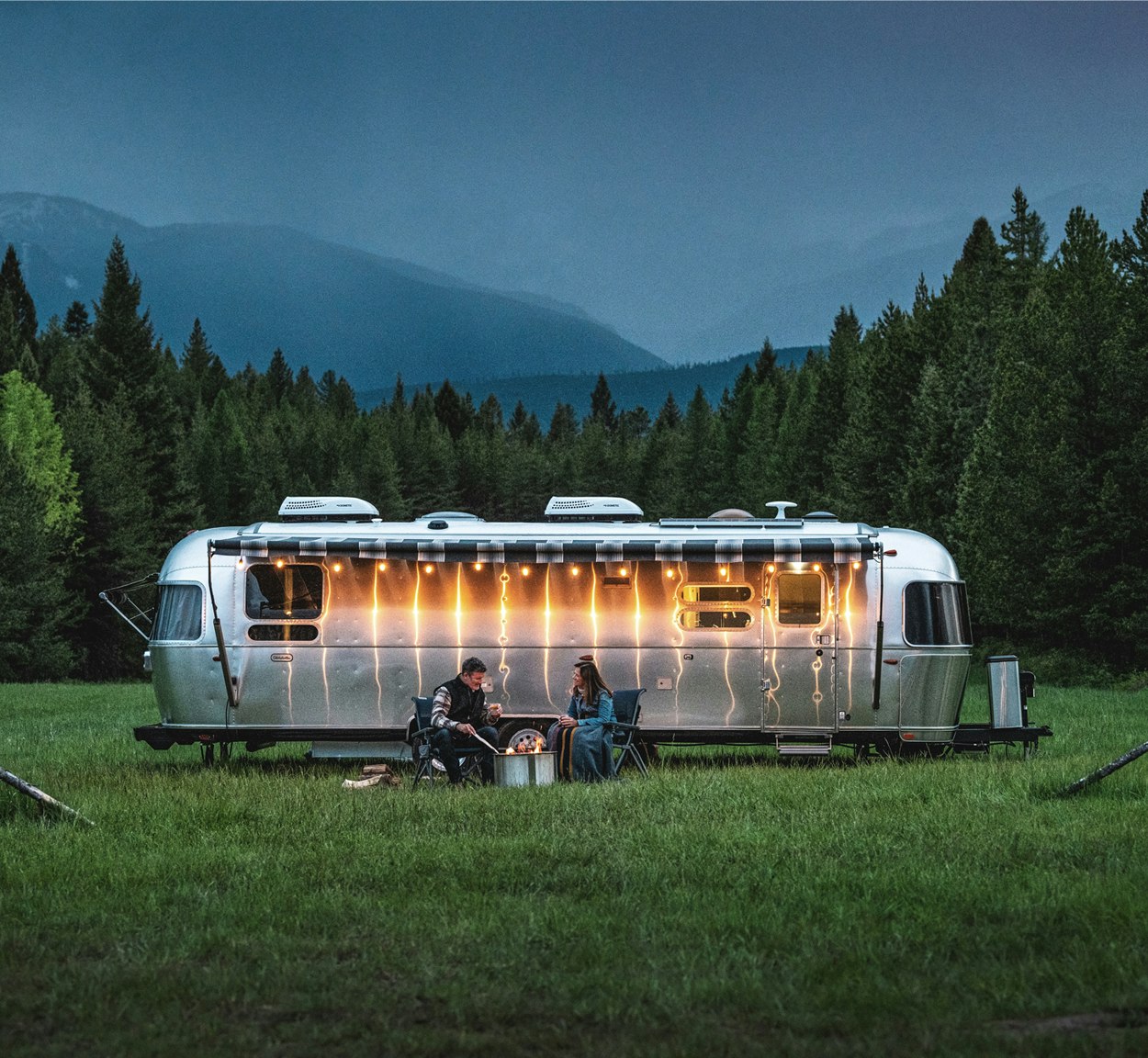 An Airstream Globetrotter Travel Trailer parked out a park with two campers sitting by a fire under the awning